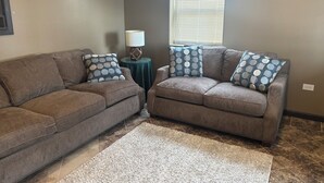 Ein Wohnzimmer mit zwei braunen Sofas, Kissen mit Mustern, einem kleinen runden Tisch mit Lampe und einem Fenster mit Jalousien.