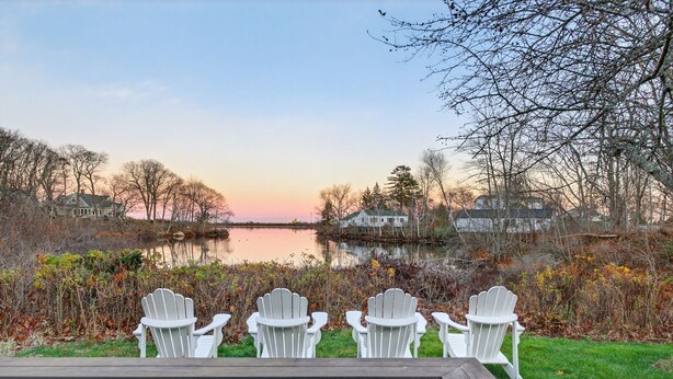 Eine Terrasse mit vier weißen Adirondack-Stühlen, die auf einen See bei Sonnenuntergang blicken.