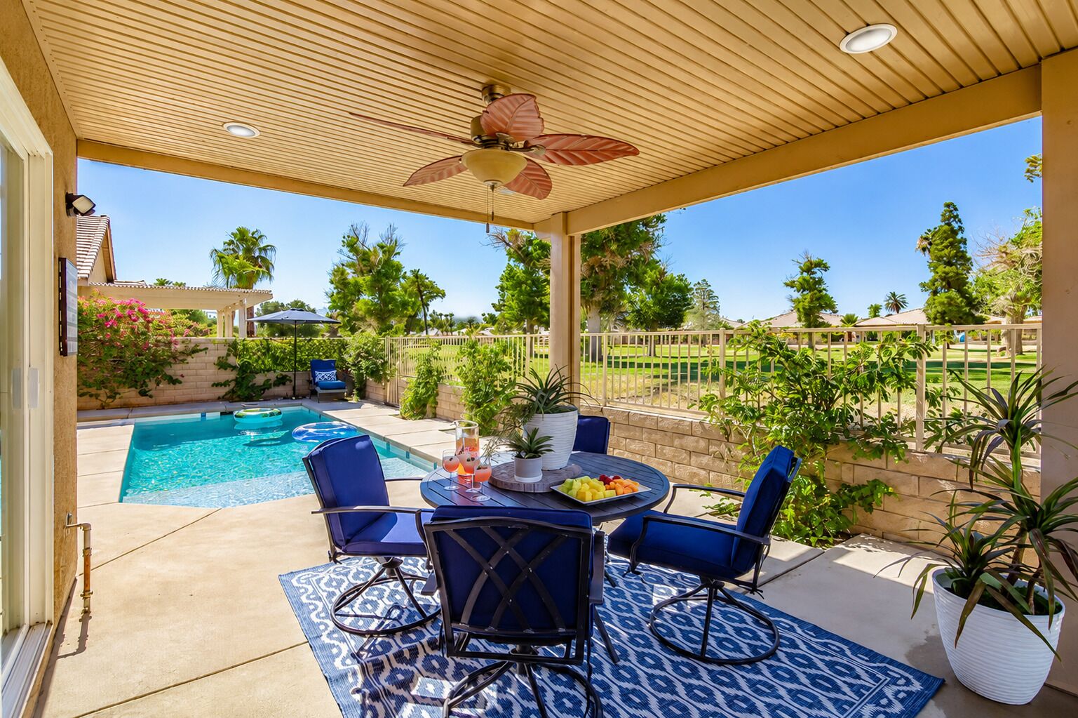 A covered patio with a pool, blue chairs, a table with fruit and drinks, and a ceiling fan.