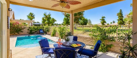 A covered patio with a pool, blue chairs, a table with fruit and drinks, and a ceiling fan.