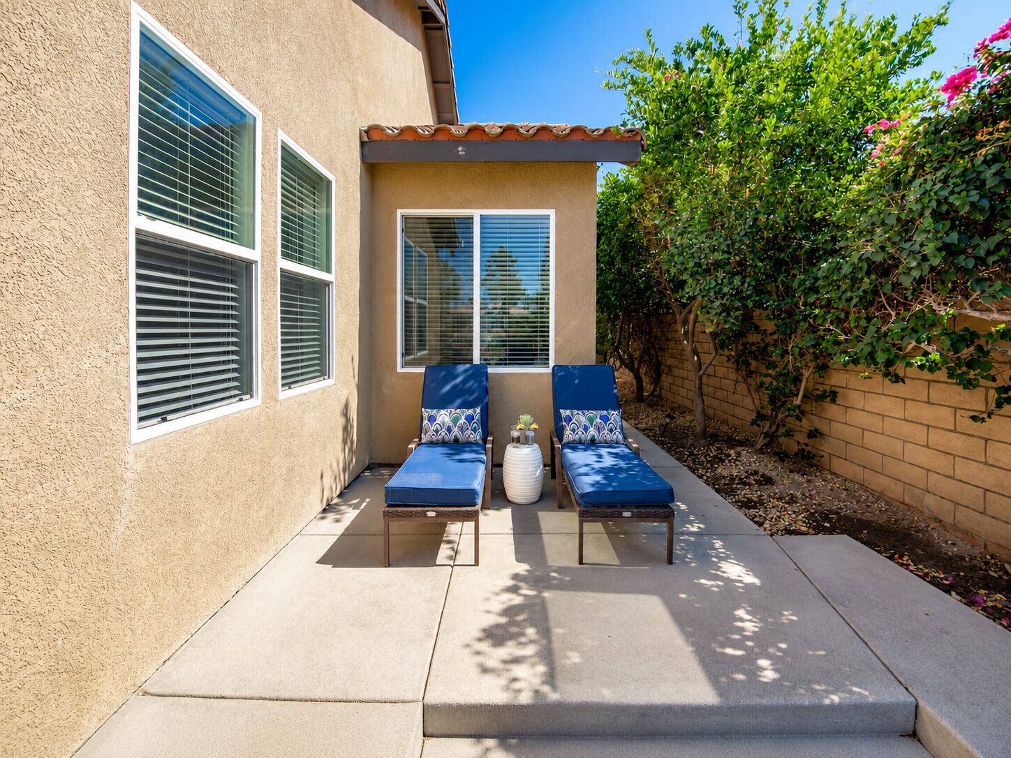 A patio area with two lounge chairs and a small table, a window with blinds, and a brick wall with greenery.