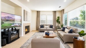 Uma sala de estar moderna com televisão de tela plana, lareira, dois sofás, uma mesa de centro e janelas grandes com persianas.