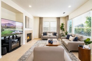 Ein modernes Wohnzimmer mit Flachbildfernseher, Kamin, zwei Sofas, einem Couchtisch und großen Fenstern mit Jalousien.