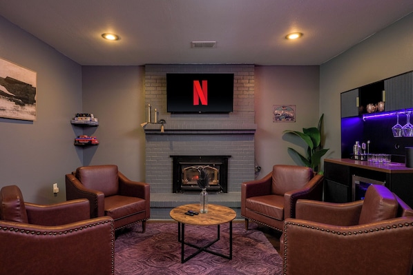 A cozy living room with leather armchairs, a fireplace, a mounted TV, and a bar area with blue lighting.