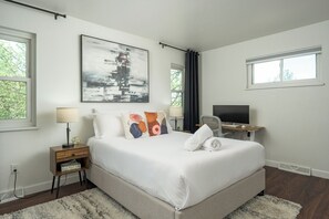 A bedroom with a large bed, a desk with a computer, a window with curtains, and a painting on the wall.