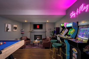A room with a pool table, two leather chairs, a fireplace, a neon sign, and arcade games.