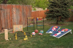 A backyard with a wooden fence, a tree, and various outdoor games including a hopscotch board, a basketball hoop, and a seesaw.