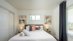 Une chambre avec un grand lit, deux tables de chevet avec des lampes, une fenêtre avec des rideaux et deux tableaux abstraits accrochés au mur.