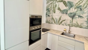 A modern kitchen with white cabinetry, a black oven, a stainless steel sink, and a decorative wall mural of tropical plants.