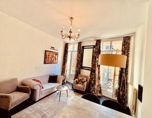 Sala de estar com sofá, poltrona e mesa. Há um candelabro, cortinas e uma janela com vista.