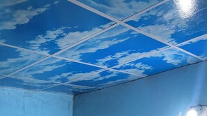 Une pièce avec un plafond bleu, une petite fenêtre munie d’une moustiquaire, une boîte électrique fixée au mur et une moquette verte sur laquelle repose un tapis à motifs.