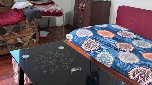 Een kleine, goed verlichte kamer met een bed, een houten tafel, een stoel, een klein kastje en een raam.