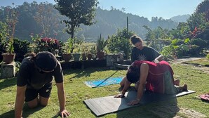 Twee personen doen yoga op matten buiten, met een heldere lucht en een boom op de achtergrond.