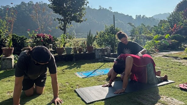 Deux personnes pratiquent le yoga sur des tapis à l’extérieur, sous un ciel dégagé, avec un arbre en arrière-plan.