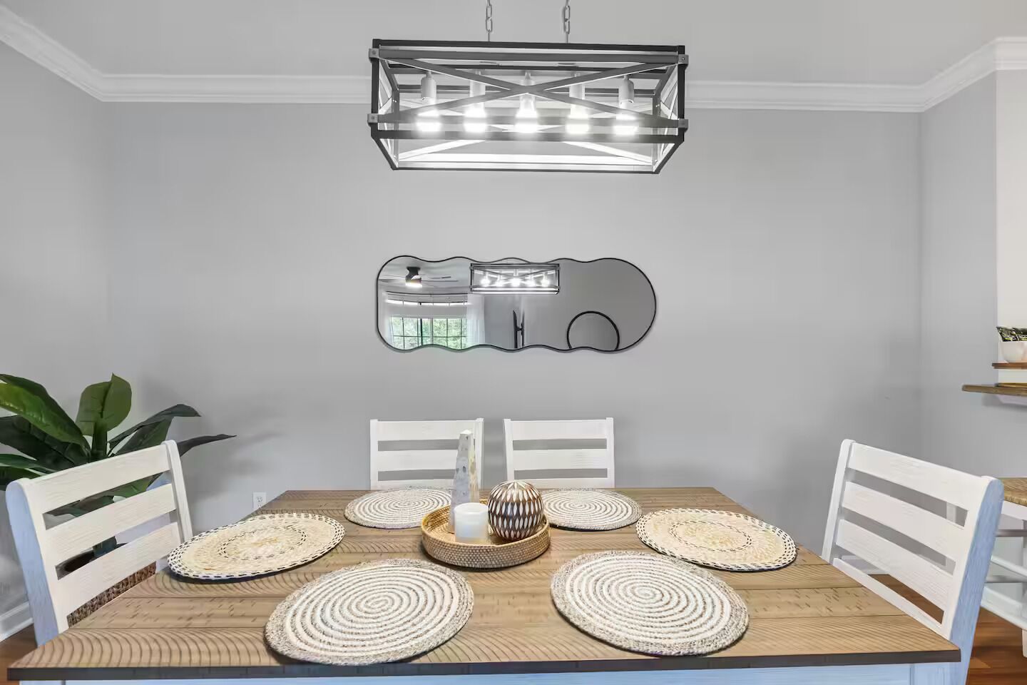 Un coin repas avec une table en bois, des chaises blanches, des sets de table en fibres tressées, un chandelier central et une suspension moderne.