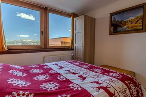 Ein Schlafzimmer mit einem großen Fenster, einem Bett mit einer rot-weißen Steudecke, einem Heizkörper, einem Holzschrank und einem gerahmten Landschaftsbild an der Wand.