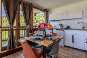 Ein Essbereich mit Tisch für vier Personen, eine Küche mit weißen Schränken und Blick auf eine bewaldete Landschaft durch große Fenster.