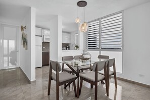 Un espace repas moderne avec une table en verre et des chaises en bois, une cuisine en arrière-plan et une grande fenêtre avec des stores.