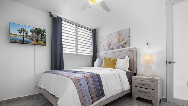 A bedroom with a bed, a nightstand, a lamp, a painting, and a window with blinds. - Designer remodeleded apt.View, Parking, location,NEW listing! (Luquillo)