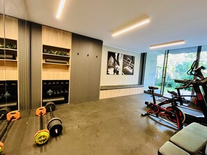 A modern gym with weightlifting equipment, a stationary bike, and a large window offering a view of the outdoors. - Modern Studio Las Tranqueras - Pool, Gym & Laundry (Las Condes)