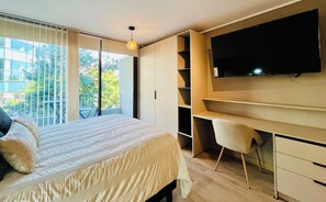 A bedroom with a bed, a TV mounted on the wall, a desk with a chair, and a balcony with a view of trees. - Modern Studio Las Tranqueras - Pool, Gym & Laundry (Las Condes)