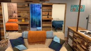 Een gezellige kamer met een bank, twee bedden, een houten wand, een decoratieve spiegel en een kitchenette.