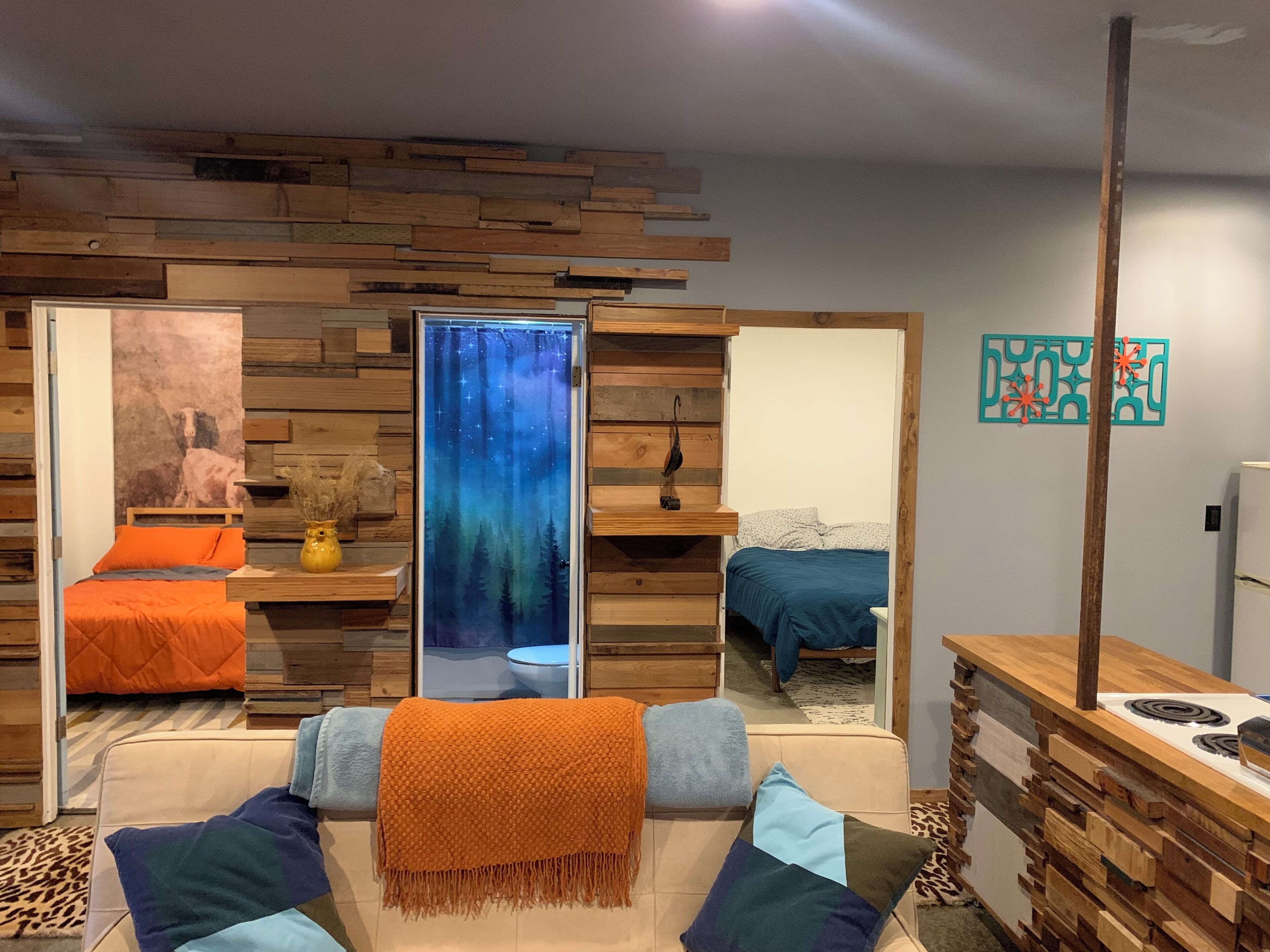 Ein gemütliches Zimmer mit einer Holzwand, einem Bett mit orangefarbener Bettwäsche, einem Sofa mit blau-orangenen Kissen und einem Fenster mit Sternenhimmel-Motiv.