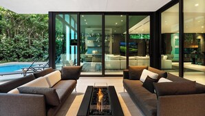 A modern living room with a glass fireplace, comfortable seating, and a view of a pool and greenery. - Villa Strata: Modern 5 BED +POOL, minutes from The Grove, Brickell & Downtown (Miami)