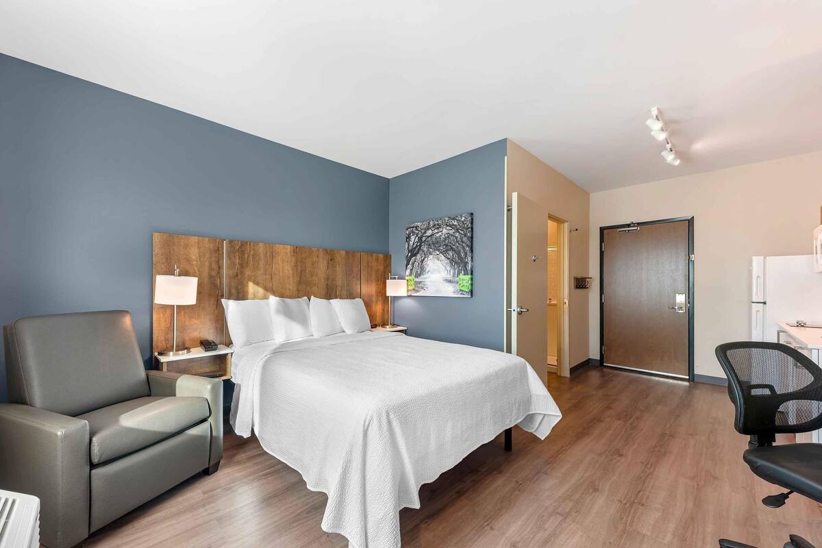 A hotel room with a bed, a chair, a desk, and a door.