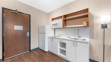 A compact kitchen with white cabinets, a microwave, a refrigerator, and a wooden shelf above the countertop.