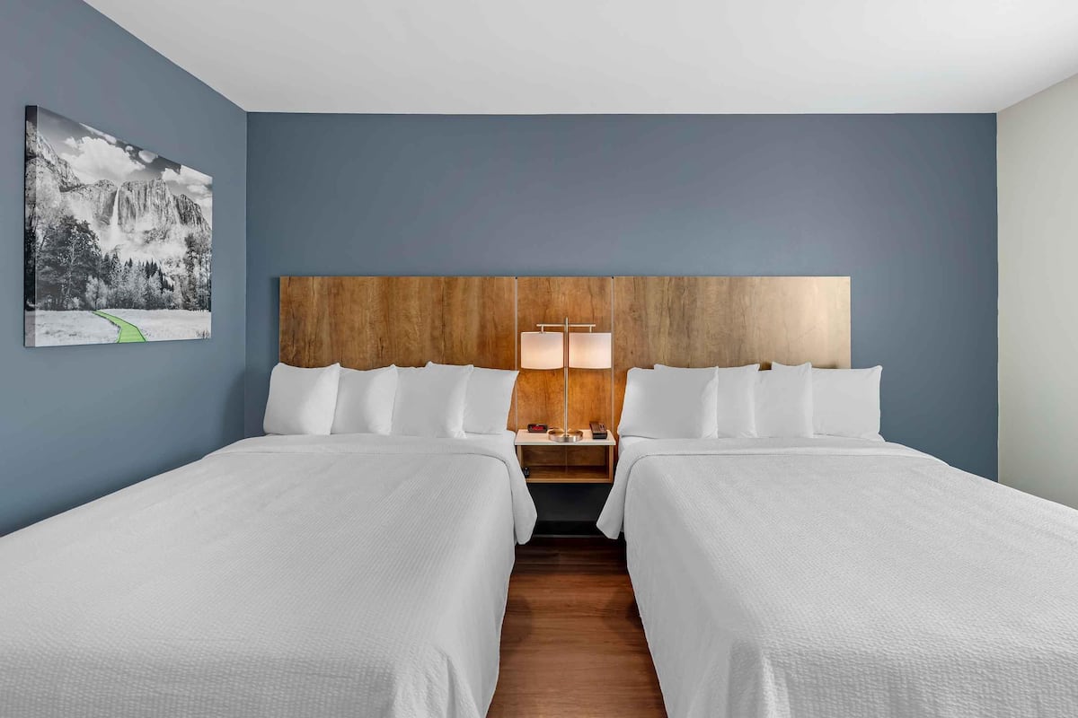 A hotel room with two beds, a wooden headboard, a nightstand with a lamp, and a framed picture on the wall.