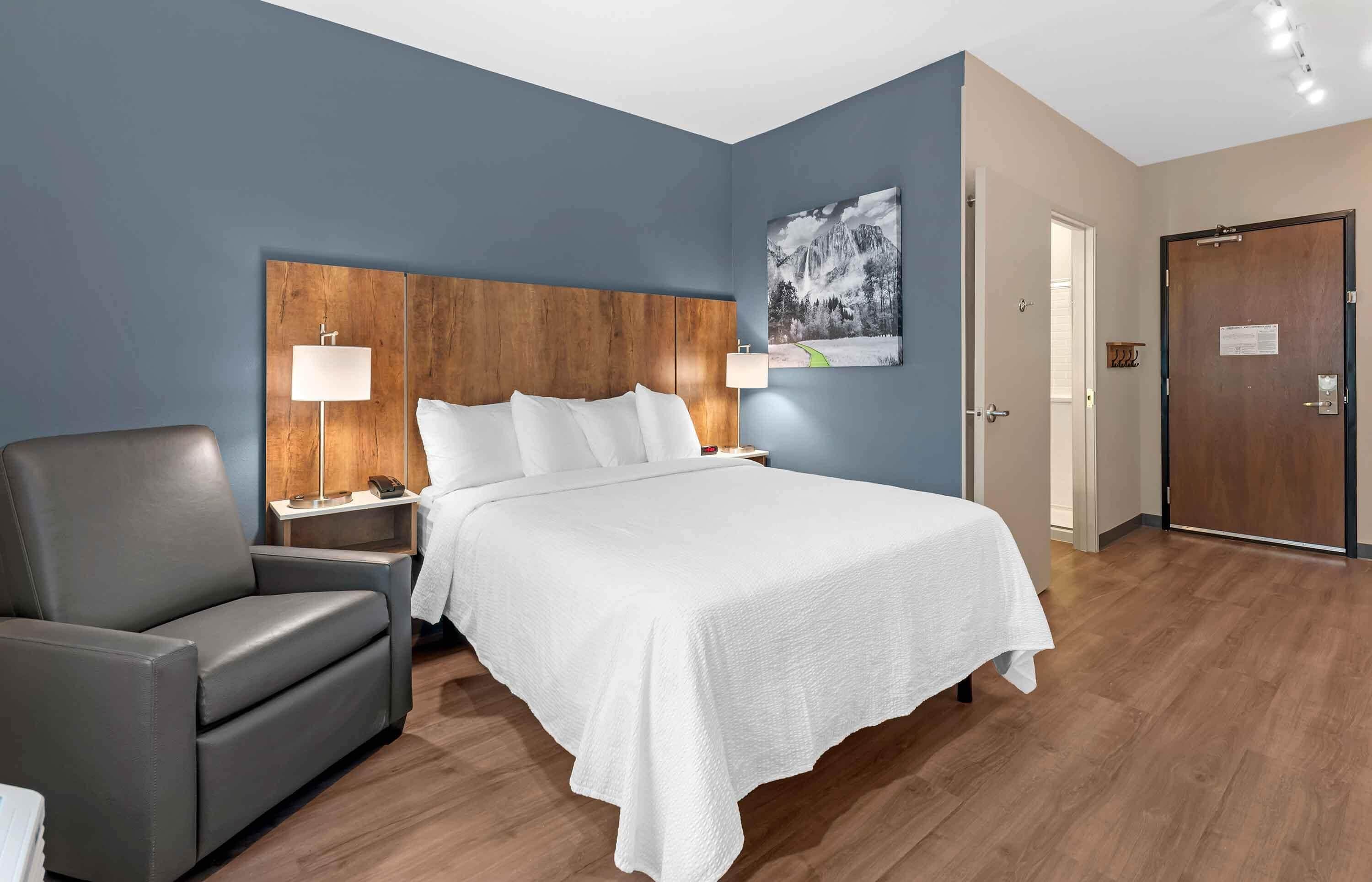 Una habitación de hotel con cama, una silla, mesita de noche, lámpara y puerta.