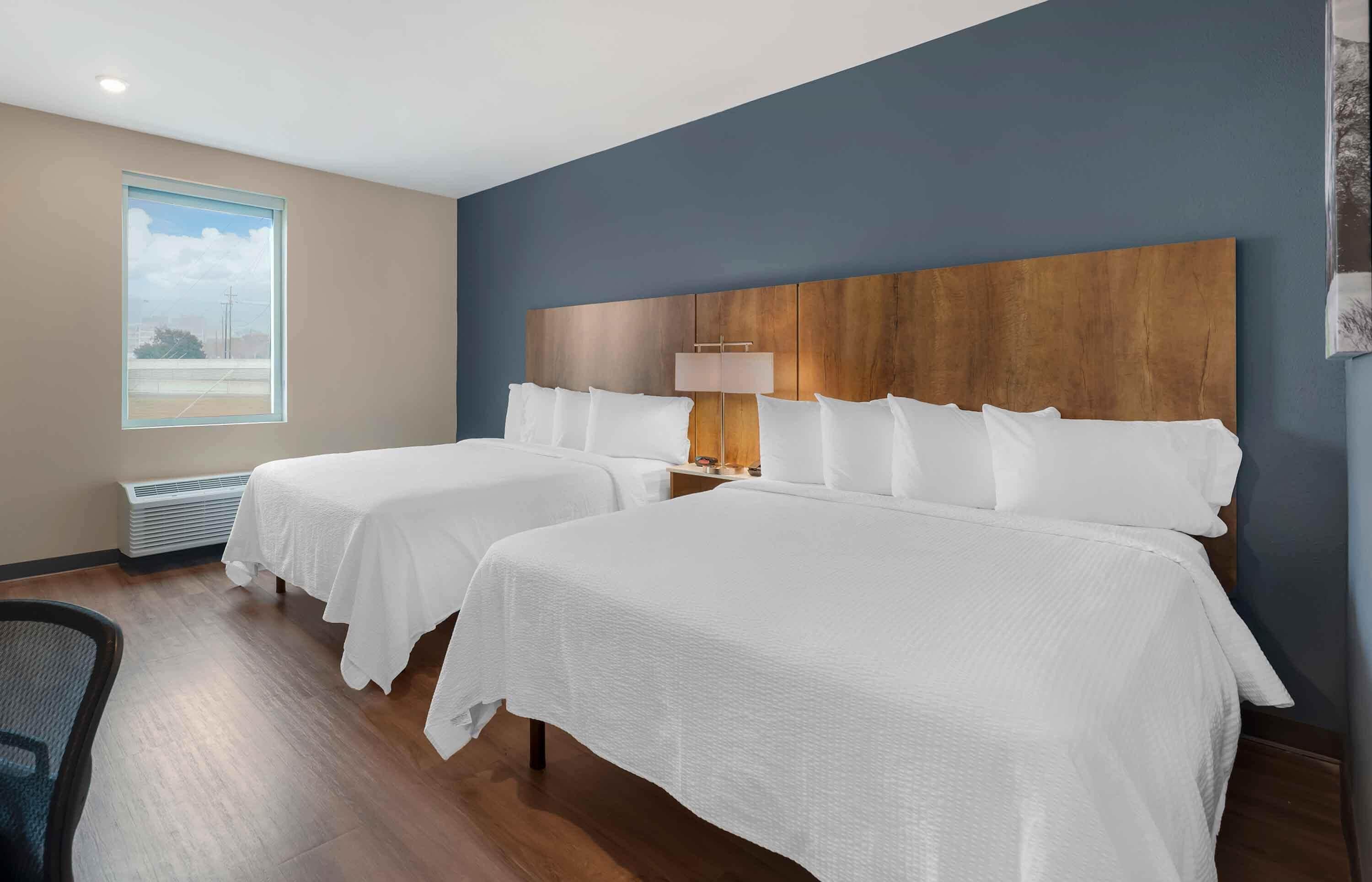 Une chambre d’hôtel avec deux lits, une grande fenêtre, une tête de lit en bois et une lampe de chevet.