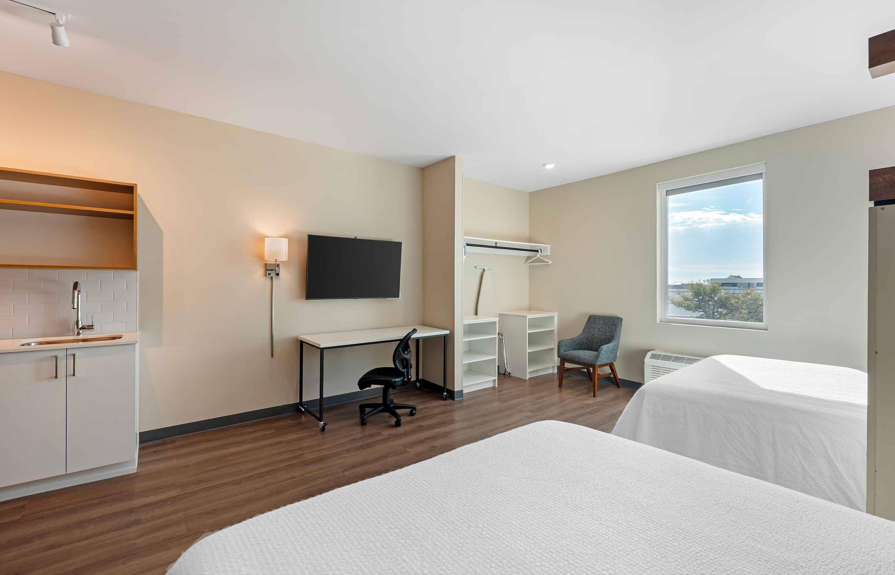 Een hotelkamer met een kitchenette, een bureau met stoel, een televisie, een kledingkast, een bed en een raam met uitzicht.