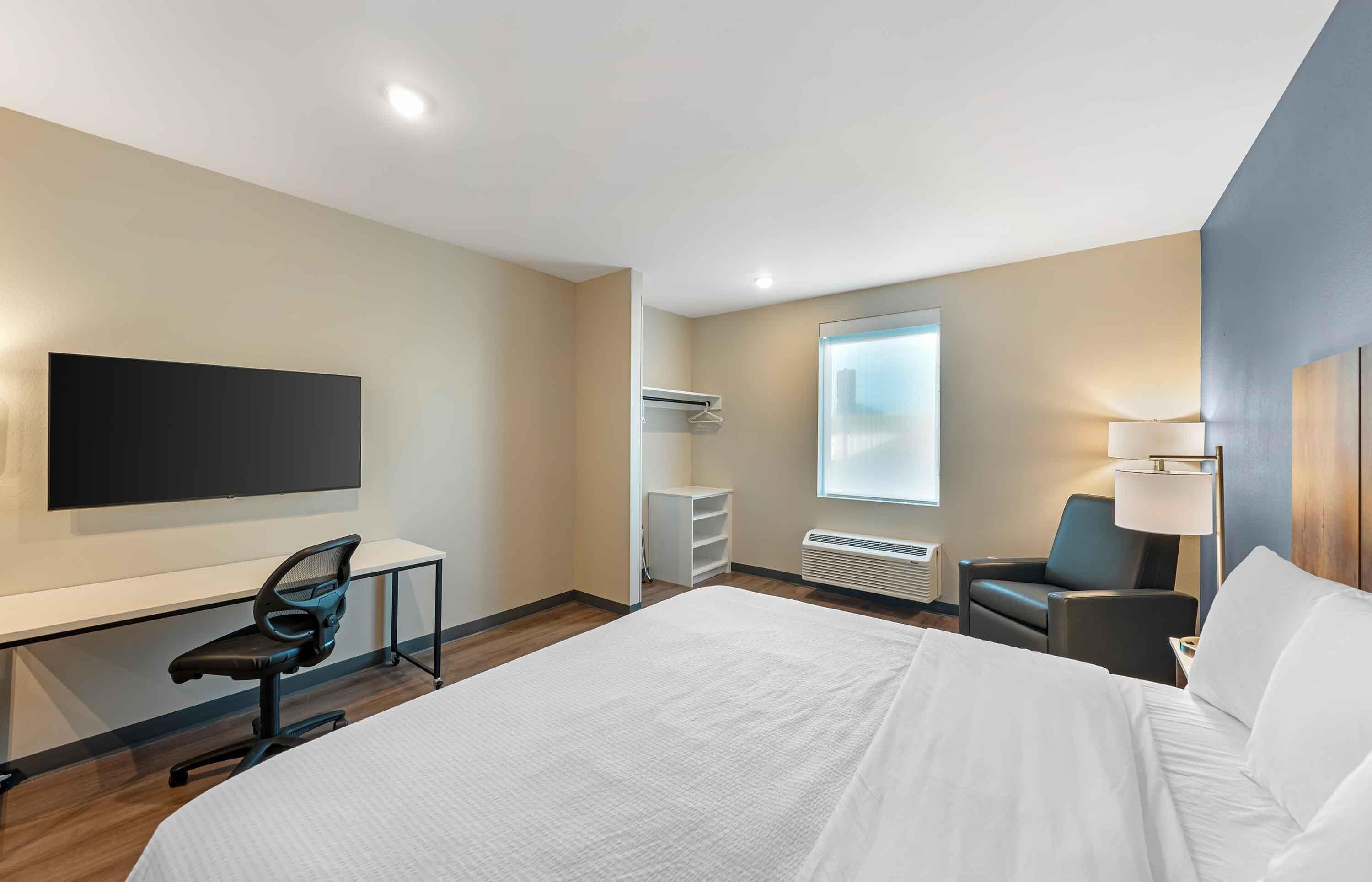 A hotel room with a large bed, a desk with a chair, a TV mounted on the wall, a window with a view, and a lamp.