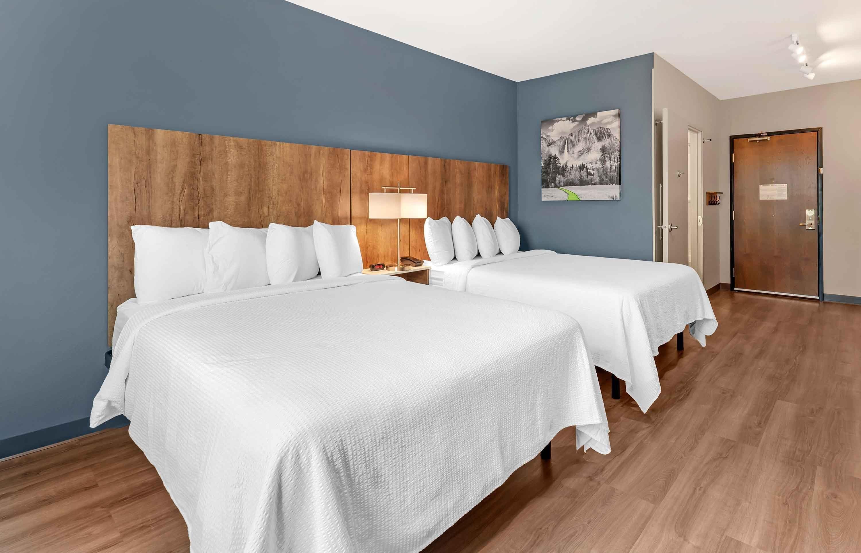 Quarto de hotel com duas camas, cabeceira de madeira, roupa de cama branca, mesa de cabeceira com abajur e porta com aviso.