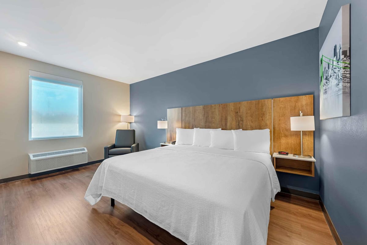 A modern hotel room with a large bed, a chair, a nightstand with a lamp, and a window with a view.