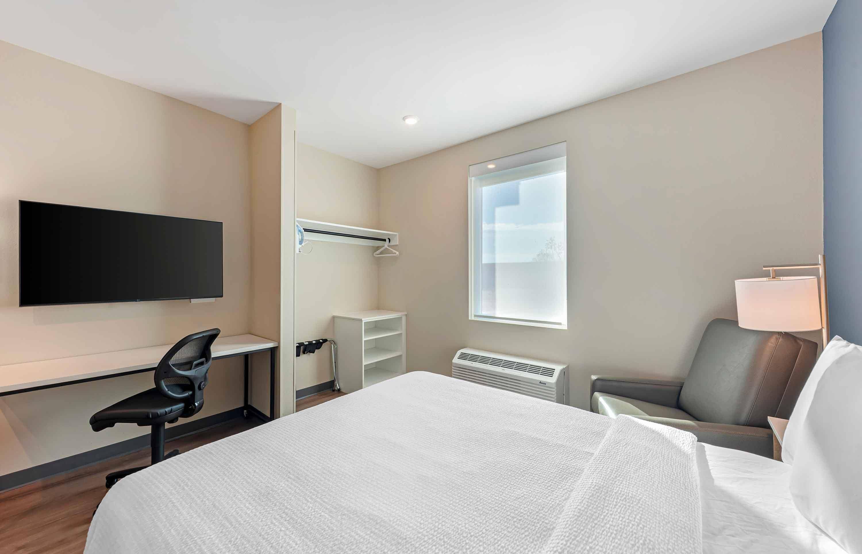 A hotel room with a bed, a desk, a chair, a TV mounted on the wall, and a window.