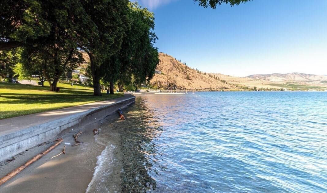 A lakeside park with a walking path, trees, and a view of hills in the distance.