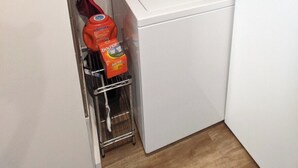 A compact laundry room with a front-loading washer and dryer, a metal rack with cleaning supplies, and a broom leaning against the wall. - Romantic Boutique Getaway with Modern Remodel (Chelan)