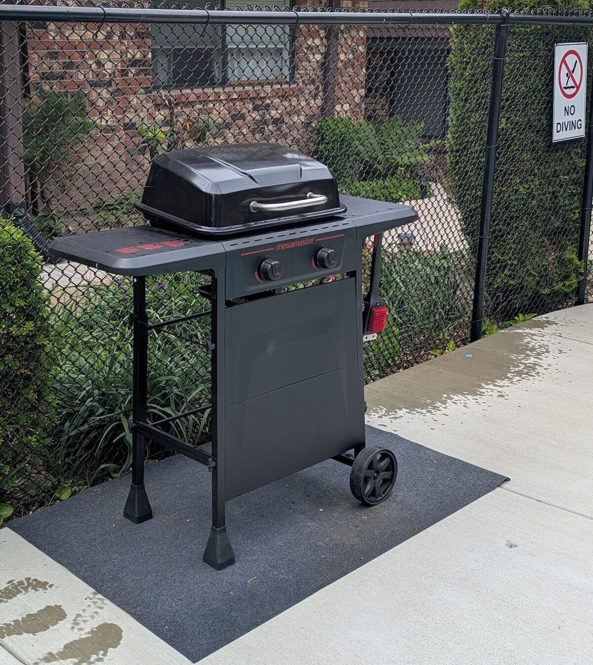 A portable barbecue grill with wheels, placed on a mat near a chain-link fence.