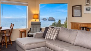 Ein Wohnzimmer mit Sofa, Sessel, Tisch und Lampe. Zwei große Fenster bieten Blick auf das Meer und Felsen.