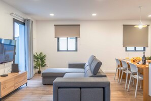 Un salon moderne avec un canapé gris, un coin repas comprenant une table et des chaises en bois, et un téléviseur posé sur un meuble en bois.