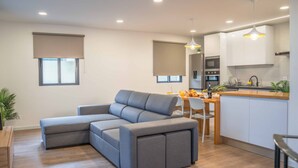 A modern living room with a grey sectional sofa, a dining area with a wooden table and chairs, and a kitchen with white cabinets and a built-in oven. - A View to Paradise – Spectacular Sea & Bay Views (Funchal)