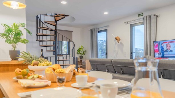 A breakfast setup with fruit, bread, and orange juice on a wooden table, a grey sofa, a spiral staircase, and a TV displaying a news program. - A View to Paradise – Spectacular Sea & Bay Views (Funchal)