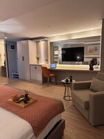 Una habitación de hotel moderna con una cama grande, un sofá, un escritorio con una computadora portátil, un televisor y una zona de kitchenette.