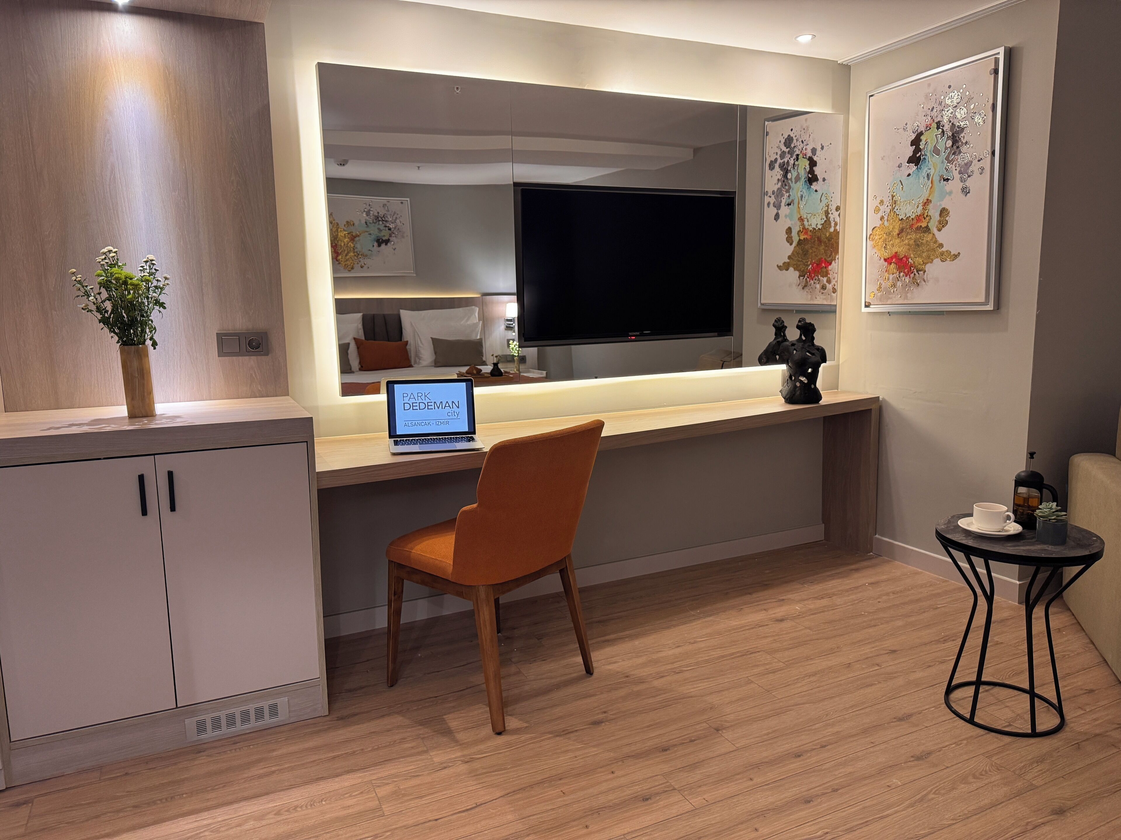 A modern hotel room with a wooden desk, a laptop, a chair, a small table with a cup and a plant, and artwork on the walls.