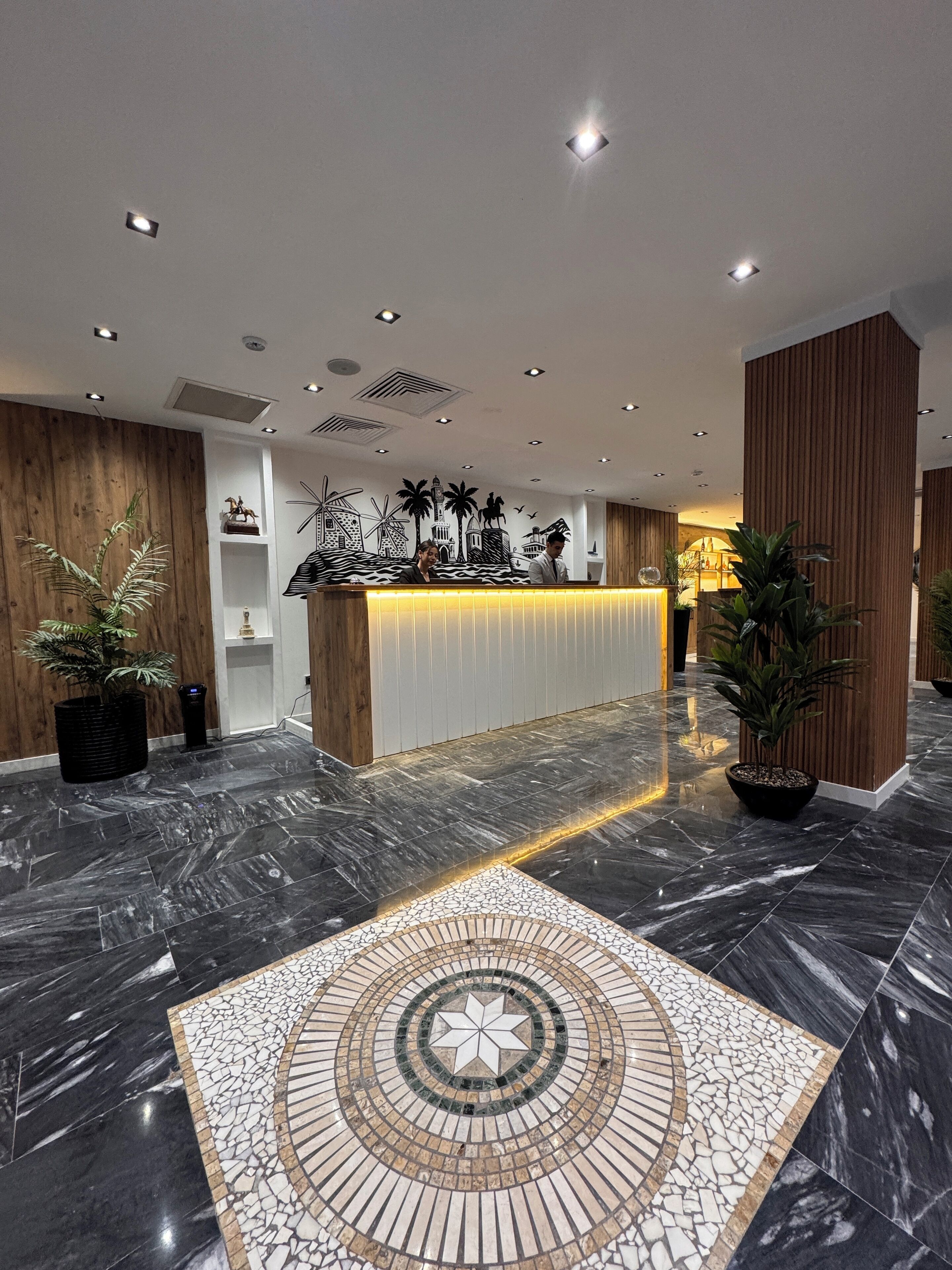 A hotel reception area with a modern design, featuring a marble floor, a central mosaic pattern, wooden paneling, and a reception desk with a decorative backdrop.