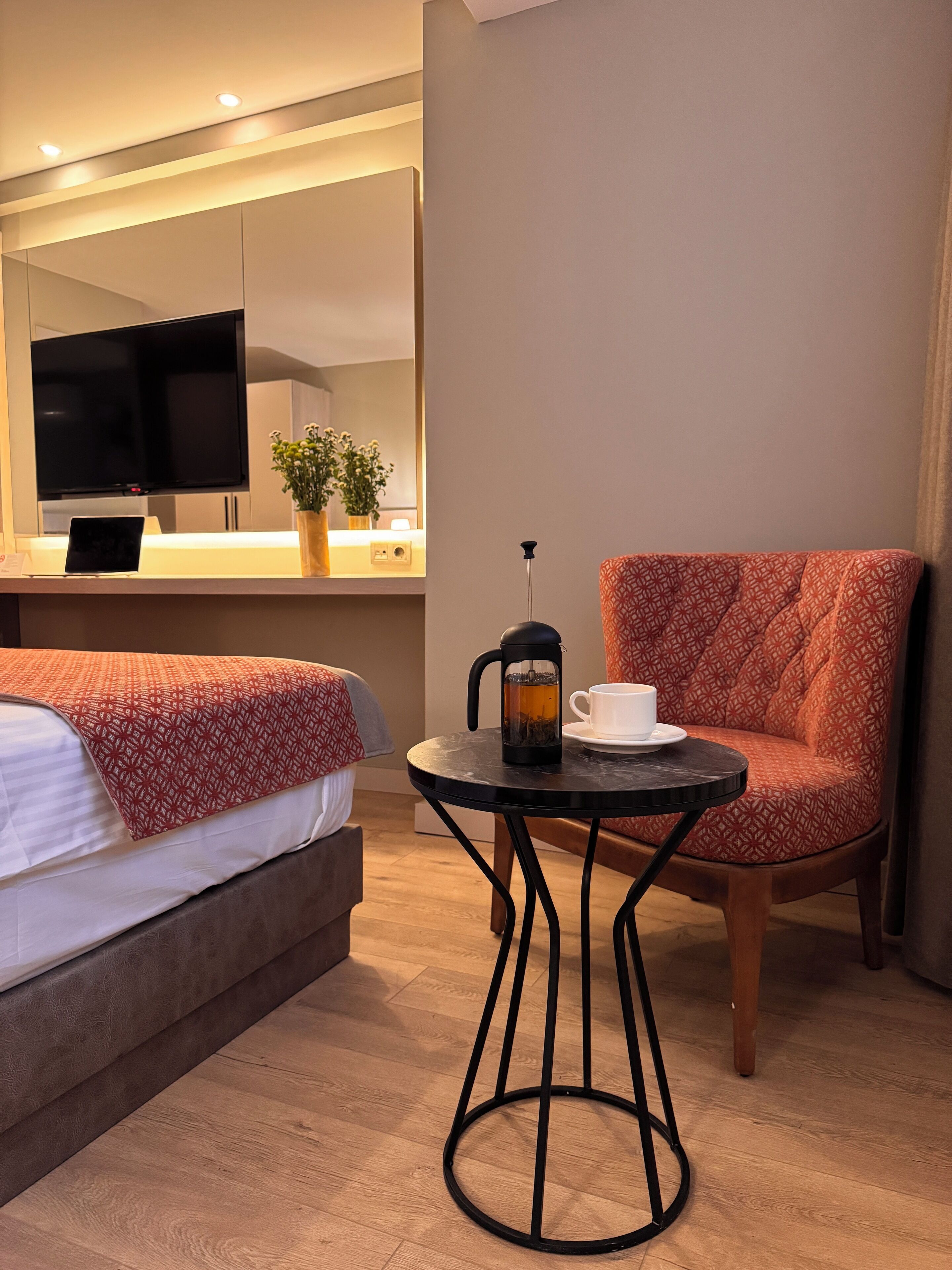 Habitación de hotel con cama, una mesita con cafetera y taza, una silla, televisor y planta.