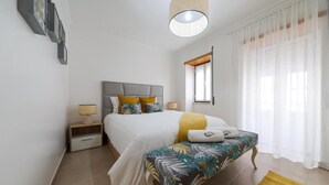 Une chambre à coucher comprenant un lit, des tables de chevet, un banc, un luminaire suspendu et une fenêtre avec des rideaux.